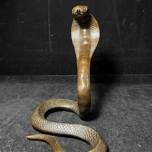 May include: A bronze-colored metal cobra figurine in an upright, alert posture with its hood flared. The sculpture features a detailed, textured scale pattern along its coiled body and a smooth, vertical ribbed design on the front of its hood.