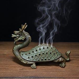 Könnte beinhalten: Ein bronzefarbener Räucherstäbchenhalter aus Metall in Form einer Fabelwesen-Drachenschildkröte. Der Panzer besteht aus überlappenden kreisförmigen Münzmustern mit kleinen quadratischen Aussparungen, aus denen dünne weiße Rauchschwaden aufsteigen.