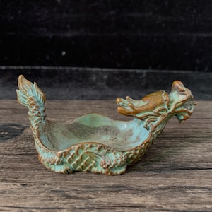 May include: A small, open-top bowl shaped like a dragon, featuring a weathered, verdigris-toned patina over a metallic base. The dragon's head extends from one side, while its tail curves upward on the other, with detailed scale textures.