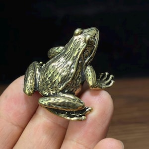 以下が含まれることがあります： 表面に凹凸のある質感を持つ、金色の金属製カエルの置物。後ろ足を折り曲げ、前足を伸ばした座った姿勢で、目や水かきのある指先など、詳細な解剖学的特徴が表現されています。