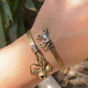 Könnte beinhalten: Ein messingfarbenes Metallarmband mit einer Pferdefigur, die mit kleinen klaren Steinen verziert ist. Das Armband enthält einen herzförmigen Anhänger und zwei kleine runde Glöckchen, die mit einem gewickelten Metalldetail am Band befestigt sind.