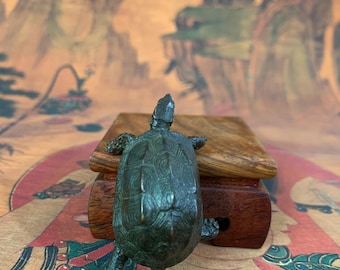 May include: A bronze turtle figurine with detailed shell patterns sits atop a small, wooden box. The turtle is positioned facing away from the viewer, showcasing its textured shell and legs. The background features a colorful, patterned surface.
