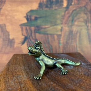 May include: A brass figurine of a smiling lizard with a spiky crest. The lizard is posed on a wooden surface, with a detailed, textured appearance. The background features a landscape painting with earthy tones.