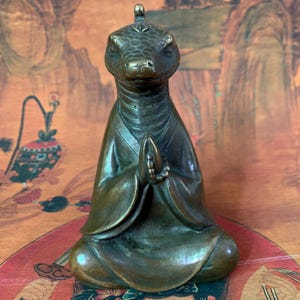 May include: Bronze sculpture of a meditating reptile-like figure in a seated position with hands clasped in prayer. The figure wears a robe and has a detailed reptilian head with closed eyes. The sculpture has a dark, aged patina.