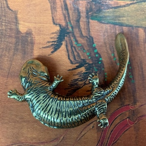 May include: A detailed bronze-colored lizard figurine with striped markings. The lizard is lying on its belly with its legs outstretched. The background features a brown and green painted design.