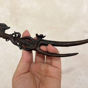 May include: A dark brown, carved wooden hair fork featuring an intricate design of a stylised animal head at one end, followed by floral motifs and swirling patterns that transition into two long, tapered prongs for securing hair.