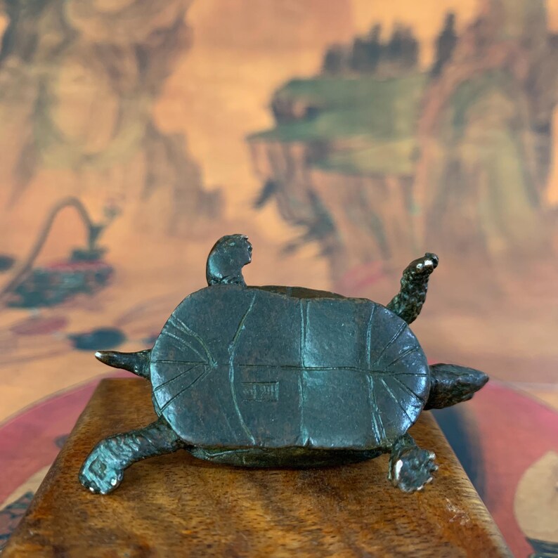 May include: A dark bronze turtle figurine with its underside visible, showcasing intricate details. The turtle is positioned on a wooden surface, with its legs and head extended. The background features a blurred landscape painting.