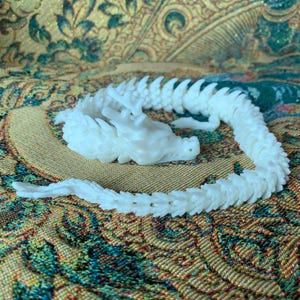 May include: A white, articulated dragon figurine with detailed scales and a flowing tail. The dragon is posed in a curved position, showcasing its intricate design. The figurine is set against a patterned background.