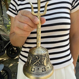Puede incluir: Una campana de mano de metal color latón con un mango de tres puntas coronado por un símbolo yin-yang. El cuerpo de la campana está decorado con intrincados relieves de motivos tradicionales, incluyendo nubes y patrones geométricos.
