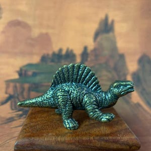 Può includere: Statuetta dettagliata di dinosauro Spinosaurus color bronzo. La statuetta ha una superficie strutturata e una grande vela sulla schiena. È posizionata su un piccolo blocco di legno quadrato, con uno sfondo sfocato di paesaggio.