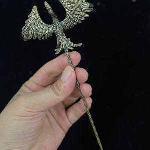 May include: A bronze-toned metal hair pin featuring a detailed phoenix ornament with outstretched wings at the top. The stem of the hair pin has a twisted, spiral design that tapers to a sharp point at the bottom end of the accessory.