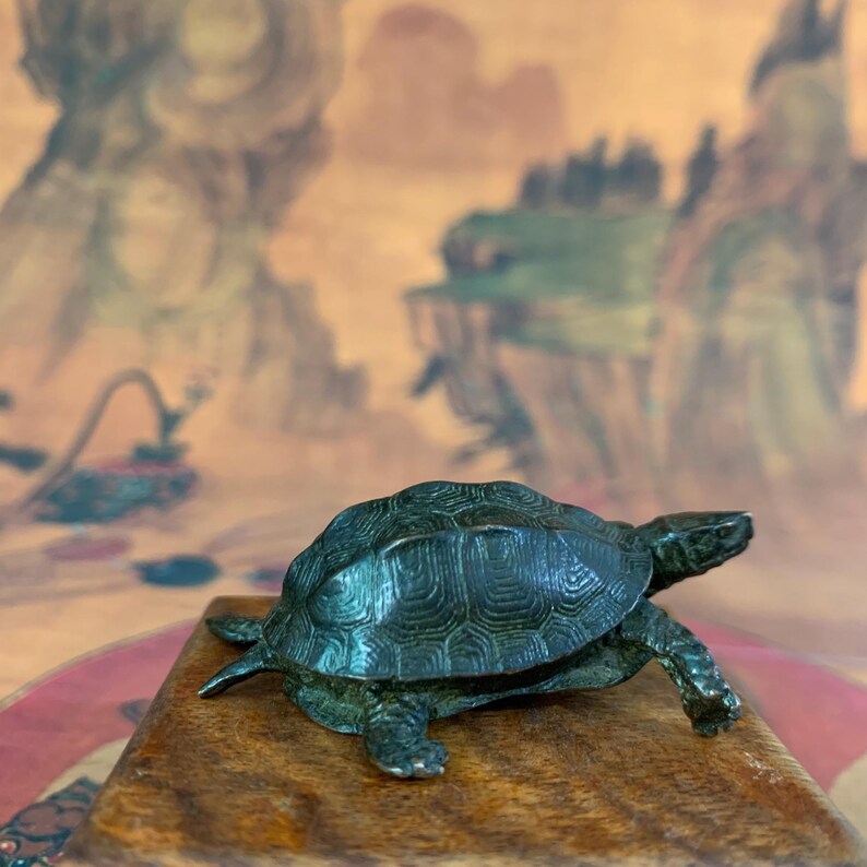 May include: A small, detailed bronze turtle figurine with a textured shell and outstretched limbs. The turtle is positioned on a wooden surface, showcasing its intricate design. The figurine is a decorative item.