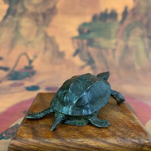 May include: A detailed bronze turtle figurine with textured shell and legs, resting on a wooden surface. The turtle is a dark green color. The background features a blurred landscape painting.