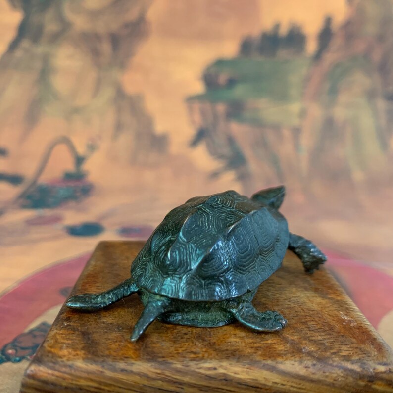 May include: A detailed bronze turtle figurine with textured shell and legs, resting on a wooden surface. The turtle is a dark green color. The background features a blurred landscape painting.