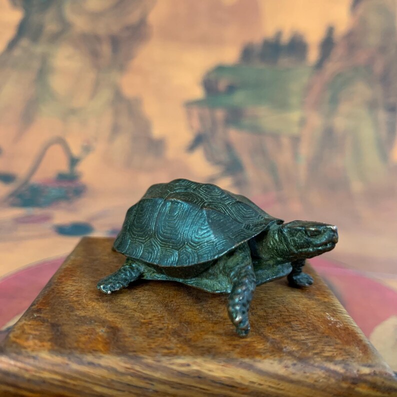 May include: A small, detailed bronze turtle figurine with a textured shell and realistic features. The turtle is positioned on a wooden base, showcasing its legs and head. The background features a blurred, artistic landscape.