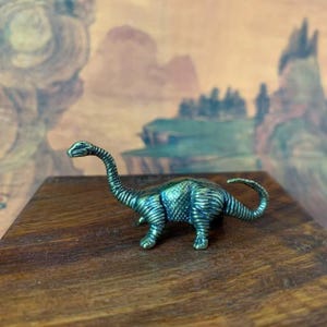 May include: A small, detailed brass dinosaur figurine with a long neck and tail. The dinosaur is a teal color with etched lines, and it is positioned on a dark wooden surface. The background features a blurred landscape.