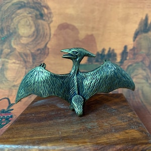Op de afbeelding: Een bronzen pterodactylusbeeldje met uitgespreide vleugels, geplaatst op een houten oppervlak. De sculptuur heeft gedetailleerde, veerachtige texturen en een kenmerkende hoofdkam. De achtergrond toont een vage, warm getinte landschap.