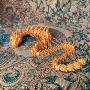 May include: A 3D-printed, articulated dragon toy in a warm orange color. The dragon features a detailed head, spiky spine, and a segmented body that allows for flexible posing. The toy is displayed on a patterned fabric.