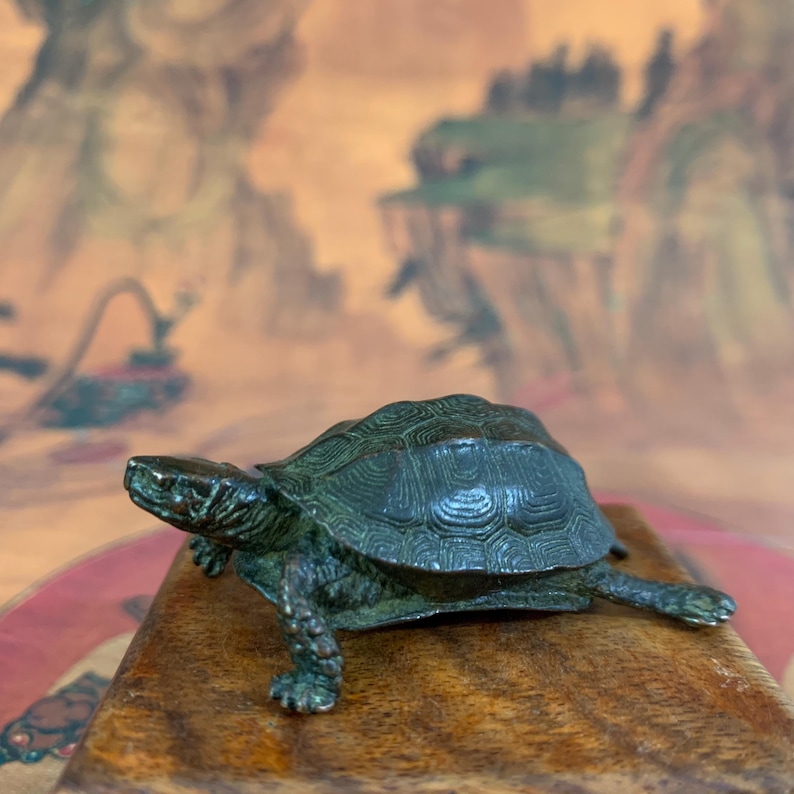 May include: A bronze-colored turtle figurine with detailed shell patterns and textured skin. The turtle is positioned on a wooden surface, showcasing its legs and head. The figurine is a decorative item.