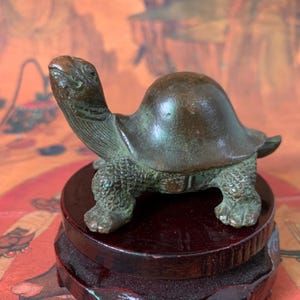 May include: A small, dark bronze-toned metal turtle figurine with a smooth, rounded shell and textured, scaly legs. The turtle is depicted with its head extended upward and is mounted on a circular, polished dark brown wooden display base.