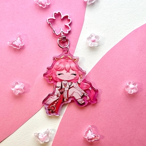 May include: A pink anime-style keychain featuring a character with pink hair and a white and red outfit. The keychain has a flower-shaped clasp. Scattered around are clear butterfly and flower-shaped embellishments on a pink and white background.