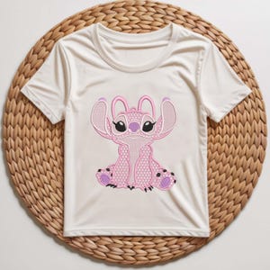 Puede incluir: Camiseta blanca de manga corta con un personaje Stitch rosa bordado. El dise&ntilde;o de Stitch incluye orejas grandes, ojos negros y detalles morados. La camiseta se muestra sobre una estera circular tejida.