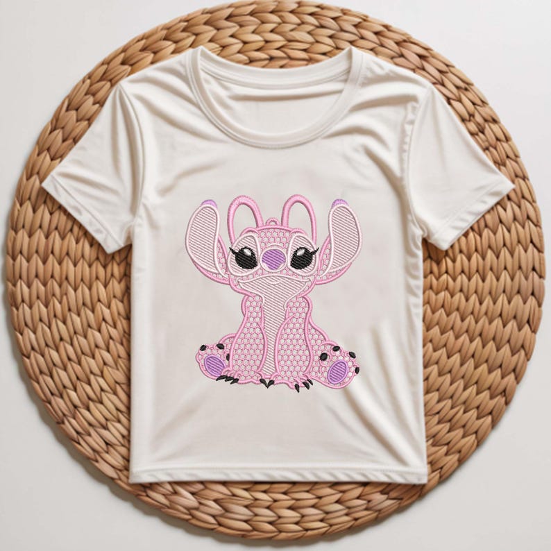 Puede incluir: Camiseta blanca de manga corta con un personaje Stitch rosa bordado. El dise&ntilde;o de Stitch incluye orejas grandes, ojos negros y detalles morados. La camiseta se muestra sobre una estera circular tejida.