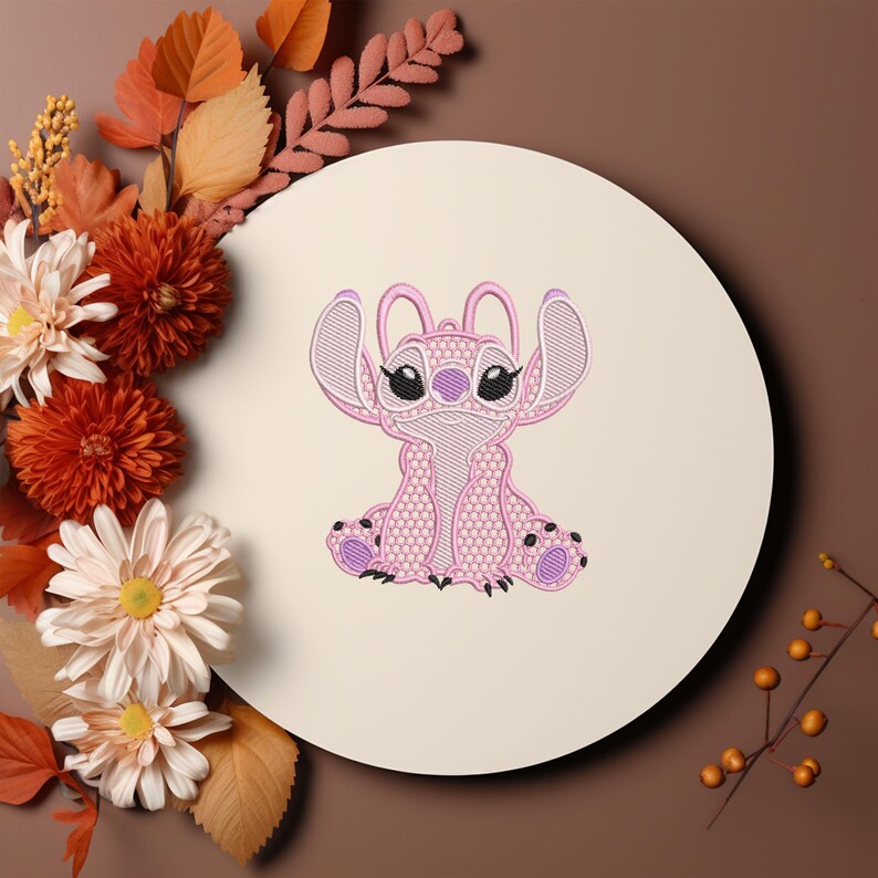 Puede incluir: Un personaje rosa bordado con orejas grandes, sentado en un c&iacute;rculo crema. El personaje tiene ojos negros y detalles morados. La imagen est&aacute; rodeada de flores y hojas oto&ntilde;ales en tonos naranja, marr&oacute;n y blanco.