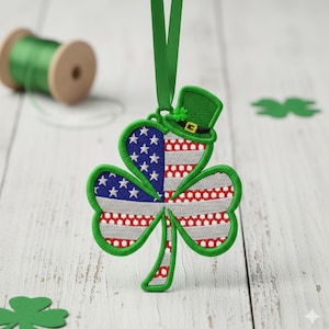May include: Embroidered shamrock ornament with a green border, featuring the American flag design on one side and a leprechaun hat. The ornament hangs from a green ribbon. A spool of green thread is in the background.