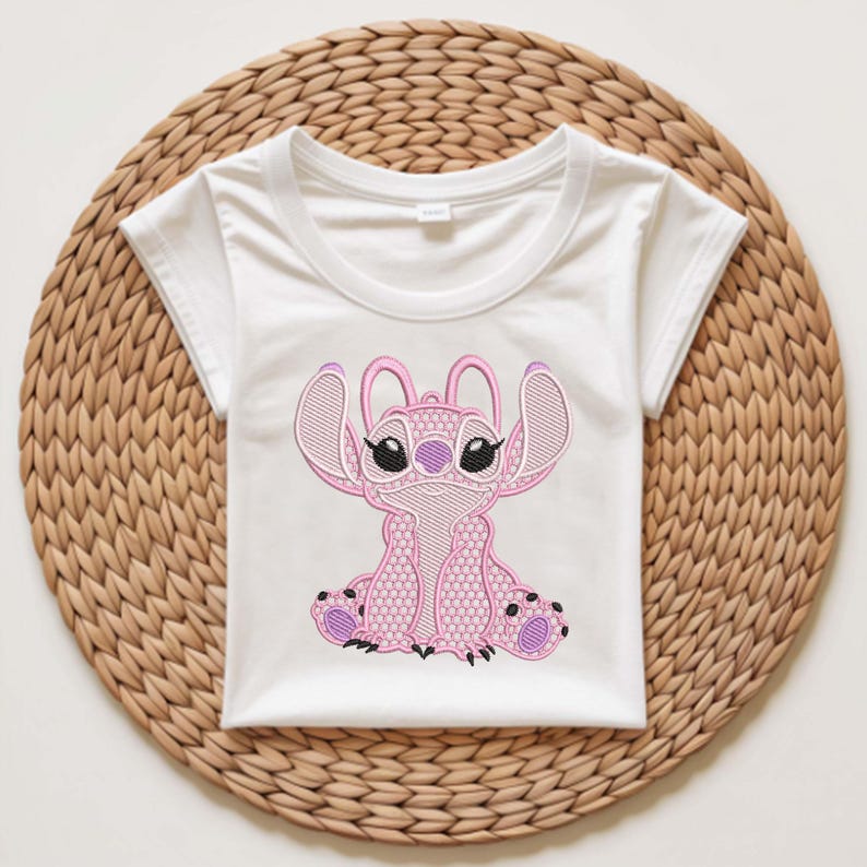 Puede incluir: Camiseta blanca con un personaje de dibujos animados rosa bordado, con grandes orejas y ojos negros. El personaje est&aacute; sentado con las piernas cruzadas. La camiseta est&aacute; sobre una estera redonda tejida.