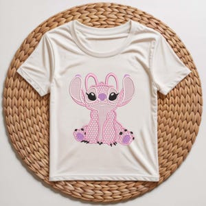 Puede incluir: Camiseta blanca con un bordado rosa del personaje Stitch. El dise&ntilde;o presenta costuras detalladas, con acentos rosas y negros. La camiseta se muestra sobre un salvamanteles redondo y tejido.