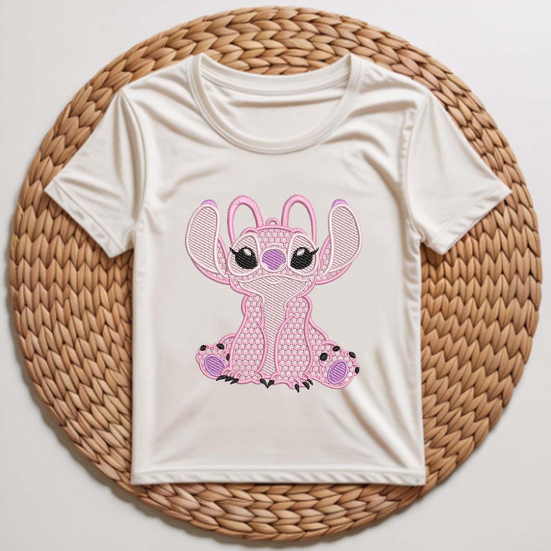 Puede incluir: Camiseta blanca con un bordado rosa del personaje Stitch. El dise&ntilde;o presenta costuras detalladas, con acentos rosas y negros. La camiseta se muestra sobre un salvamanteles redondo y tejido.