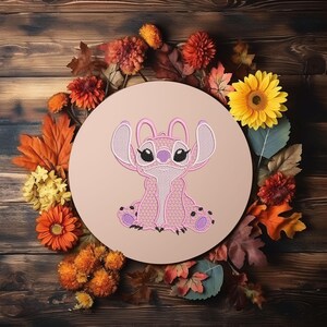 Puede incluir: Un dise&ntilde;o de personaje bordado en rosa, rodeado de hojas y flores de oto&ntilde;o, se encuentra sobre un fondo circular marr&oacute;n claro. El personaje tiene orejas grandes, ojos negros y detalles en morado. El fondo es una superficie de madera.