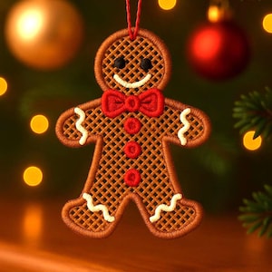 May include: Embroidered gingerbread man Christmas ornament with a red bow tie and buttons. The ornament is brown with white accents and a smiling face. It hangs from a red string against a blurred background of Christmas lights and ornaments.