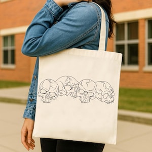 May include: Cream-colored tote bag featuring a black line drawing of four skulls. The skulls are arranged horizontally across the bag. The bag has a simple, rectangular shape with a handle.