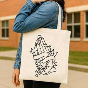 Puede incluir: Bolso tote color crema con un gráfico negro de manos rezando, un rosario y una cruz. El diseño incluye una pancarta. El bolso tiene una forma simple y rectangular con un asa.