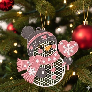 May include: A snowman Christmas ornament with a gray hat and pink scarf, holding a pink heart-shaped sign with a snowflake design. The snowman has a carrot nose and is decorated with hearts and snowflakes.