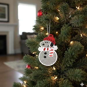 May include: A Christmas tree ornament featuring Snoopy as a snowman. The ornament has a red knit cap and scarf, with black and white details. It is hanging on a green Christmas tree with lights and other ornaments.
