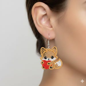 May include: Embroidered fox earrings with a red heart. The earrings feature a detailed design with brown, white, and black accents. The fox is holding a red heart. The earrings are attached to silver-colored hooks.