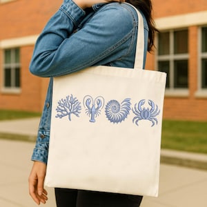May include: Cream-colored tote bag featuring blue embroidered sea life designs: coral, lobster, nautilus shell, and crab. The bag has a simple design with a rectangular shape and a handle for carrying.