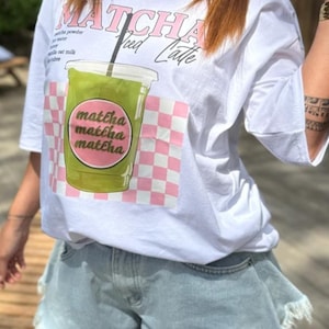 Peut inclure: Un t-shirt blanc avec un motif représentant un latte glacé au matcha vert dans un gobelet transparent. Le design inclut le texte "MATCHA Iced Latte" en rose et noir, sur un fond à carreaux rose et blanc avec le mot "matcha" répété trois fois.