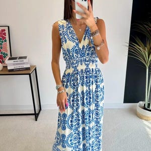 Peut inclure: Une robe longue sans manches avec une base couleur crème ornée d'un motif floral circulaire bleu vif. La robe présente un décolleté en V, une taille élastiquée froncée et une jupe fluide arrivant aux chevilles, portée avec des sandales noires.