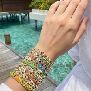 Op de afbeelding: Een stapel van negen metalen armbanden met een herhalend bloemmotief met gekleurd emaille in tinten geel, oranje, roze, groen, blauw en paars, geaccentueerd met kleine heldere steentjes in het midden van elk bloemontwerp.