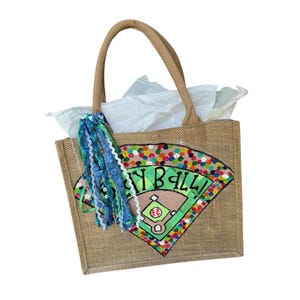 May include: A burlap tote bag with a baseball field design. The bag features a brown handle and a colorful baseball field graphic with the word "Play Ball" in green. A blue and white tassel hangs from the side.
