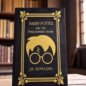 May include: A hardcover book titled "Harry Potter and the Philosopher's Stone" by J.K. Rowling. The book has a black cover with gold lettering and a gold illustration of Harry Potter's silhouette, including his glasses and lightning bolt scar. The book is on a wooden surface.