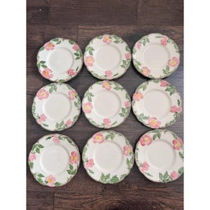 May include: Nine white ceramic plates with a floral design. Each plate features a pink rose and green leaf pattern around the rim. The plates are arranged in a 3x3 grid on a dark wood surface.