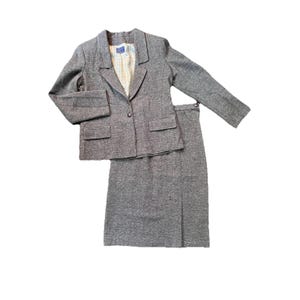 May include: A gray tweed skirt suit. The blazer has two front pockets and a single-button closure. The skirt is knee-length with a slit in the back. The suit is made of a textured fabric.
