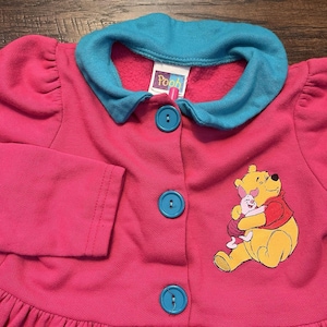 May include: A pink children's jacket with a teal collar and buttons. The jacket features an embroidered image of Winnie the Pooh and Piglet. The word "Pooh" is printed on a tag near the collar.