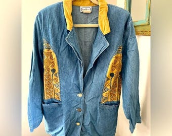 May include: Blue denim jacket with a mustard yellow collar and decorative panels. The jacket features gold-colored buttons and pockets. The decorative panels have a geometric pattern in shades of yellow, brown, and black. The jacket is hanging on a wooden hanger.