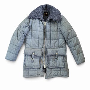May include: A light blue quilted jacket featuring a dark blue faux-fur collar and lining. The jacket has a full-length silver zipper, two large front pockets with flaps and metal hardware, and a grid-like stitched pattern throughout the fabric.